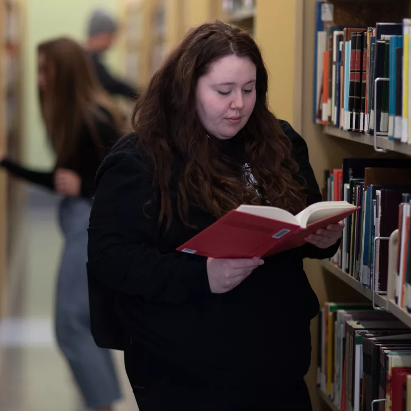 young woman looking in a book at a library, other students are looking at books on the shelves in the background